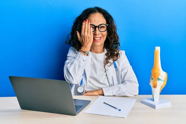 Beautiful middle age woman doctor at orthopedic clinic covering one eye with hand, confident smile on face and surprise emotion.