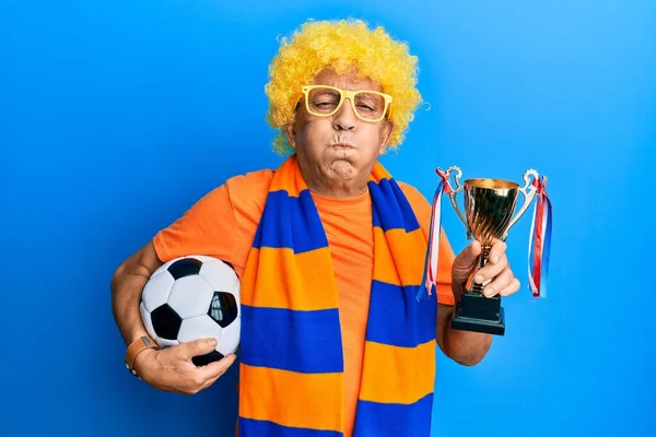 Senior hispanic man football hooligan cheering game holding ball and trophy puffing cheeks with funny face. mouth inflated with air, catching air.