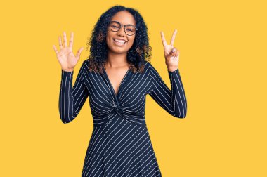 Genç Afro-Amerikalı kadın günlük kıyafetler ve gözlük takıyor. Yedi numaralı parmaklarıyla işaret ediyor. Kendinden emin ve mutlu gülümsüyor.. 