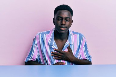 Genç, Afro-Amerikalı, gündelik giysiler giymiş, elleri karnında masada oturuyor, çünkü hazımsızlık, acı verici bir hastalık, kendini iyi hissetmiyor. Ağrı kavramı. 