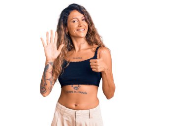 Dövmeli genç İspanyol kadın gündelik giysiler giyiyor. Altıncı parmaklarını gösterip işaret ediyor. Kendinden emin ve mutlu bir şekilde gülümsüyor.. 