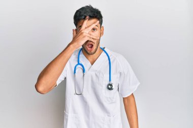Young handsome man wearing doctor uniform and stethoscope peeking in shock covering face and eyes with hand, looking through fingers afraid 