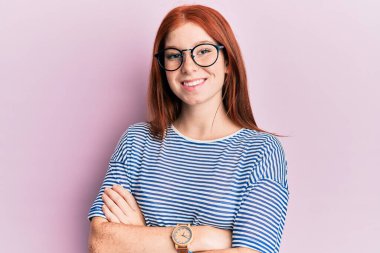 Genç kızıl saçlı, sıradan kıyafetler ve gözlüklü mutlu yüzlü, kollarını kavuşturup kameraya bakan kız. pozitif kişi. 