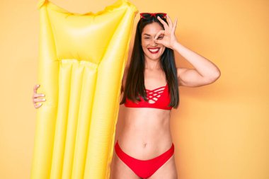 Genç, beyaz, güzel bir kadın, elinde bikinisiyle mutlu mesut gülümsüyor. 