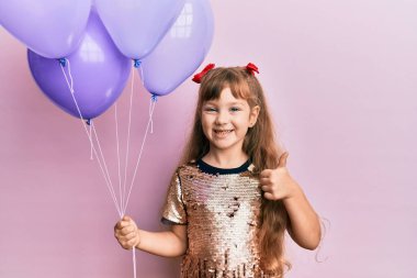 Küçük beyaz kız elinde balonlarla mutlu ve pozitif gülümsüyor, başparmağıyla mükemmel ve onay işareti yapıyor. 
