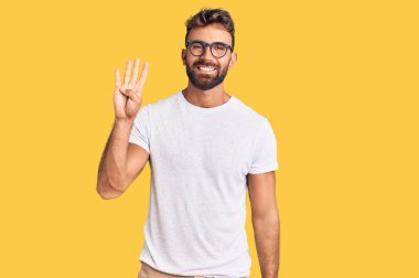 İspanyol asıllı genç adam, günlük kıyafetler ve gözlük takıyor. Dört numarayı gösteriyor ve işaret ediyor. Kendinden emin ve mutlu bir şekilde gülümsüyor.. 
