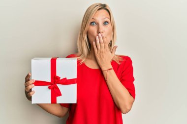 Sarışın, güzel bir kadın elinde hediyeyle ağzını eliyle kapatıyor. Şok geçiriyor ve hata yapmaktan korkuyor. şaşırmış ifade 