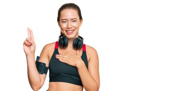 Güzel sarışın kadın spor kıyafetleri giyiyor ve kulaklık takıyor. Gülümsüyor. Elleri göğsünde ve parmakları havada küfrediyor. 