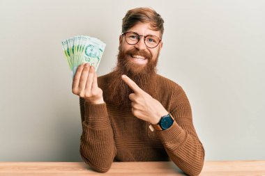 Genç İrlandalı kızıl adam elinde 5 Singapur dolar banknotlar masaya oturmuş mutlu bir şekilde el ve parmak işareti yapıyor. 
