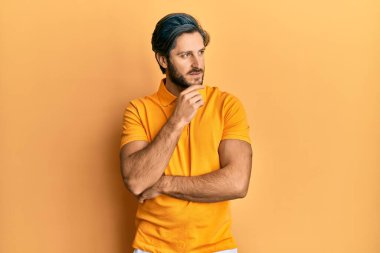 Elleri çenesinde gündelik sarı tişört giyen İspanyol genç adam soru sormayı düşünüyor, düşünceli bir ifade. Düşünceli bir yüzle gülümsüyor. şüphe kavramı. 