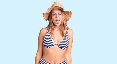 Genç, sarışın, bikini giyen ve şapkalı bir kadın kameraya seksi bir ifadeyle bakıyor. Neşeli ve mutlu bir yüzü var.. 