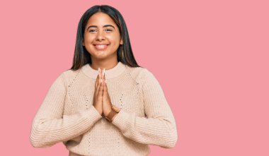 Kışlık yün kazak giyen genç Latin kız elleriyle dua ediyor af diliyor kendinden emin bir şekilde gülümsüyor.. 