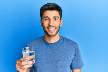 Genç yakışıklı adam bir bardak su içiyor. Pozitif ve mutlu görünüyor. Duruyor ve gülümsüyor. 
