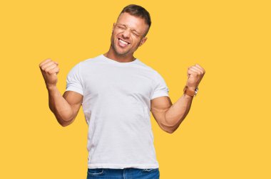 Yakışıklı, beyaz tişört giyen çok mutlu ve heyecanlı, kolları havada, gülümseyen ve başarı için bağıran, kazanan bir jest yapan kaslı adam. kutlama konsepti. 