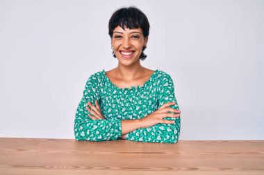 Güzel esmer kadın, sıradan kıyafetler giyip masaya oturmuş kollarını kavuşturup kameraya bakıyordu. pozitif kişi. 