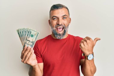 Orta yaşlı yakışıklı bir adam elinde 20 dolarlık banknotlarla baş parmağını kaldırıyor ve ağzı açık gülümsüyor. 