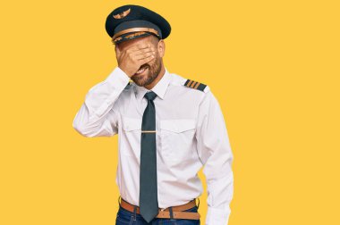 Sakallı, uçak pilotu üniforması giyen, gülümseyen ve elleri yüzünde, şaşırtmak için gözlerini kapatan yakışıklı bir adam. kör kavram. 