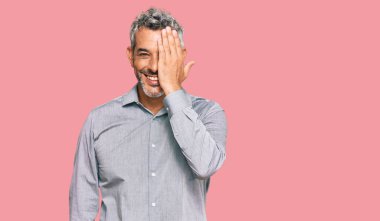 Orta yaşlı, gri saçlı, günlük kıyafetler giyen, bir gözünü elinle kapatan, yüzünde kendine güvenen bir gülümseme ve şaşırtıcı bir duygu olan bir adam.. 