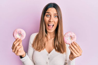 Brunette young woman holding tasty pink doughnuts celebrating crazy and amazed for success with open eyes screaming excited. 