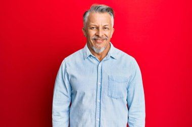 Orta yaşlı, gri saçlı, sıradan kıyafetler giyen, yüzünde mutlu ve havalı bir gülümseme olan bir adam. şanslı kişi. 