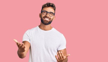 Gündelik kıyafetler ve gözlük takan genç İspanyol adam güler yüzlü ve kolları açık, dostça karşılama, olumlu ve kendinden emin selamlar. 