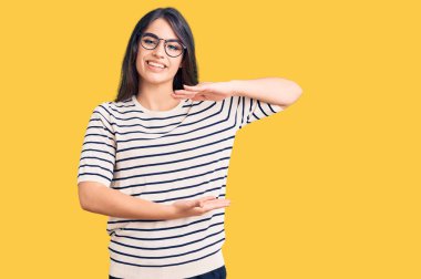 Esmer genç kız günlük kıyafetler ve gözlük takıyor. Büyük ve büyük beden işareti gösteren el kol hareketleri yapıyor. Ölçüm sembolü. Kameraya bakıp gülümsüyor. ölçüm kavramı. 