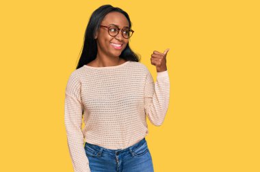 Genç siyah kadın, günlük kıyafetler ve gözlük takıyor, gülümsüyor, gülümsüyor ve başparmağıyla yan tarafa bakıyor.. 