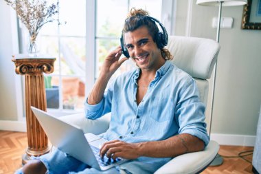 Evde dizüstü bilgisayar ve kulaklık kullanarak mutlu gülümseyen genç İspanyol adam.