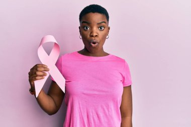 Genç Afro-Amerikalı kadın pembe kanser kurdelesi tutuyor. Korkmuş ve ağzı açık kalmış, şaşırmış ve şaşırmış. İnanmayan yüz. 