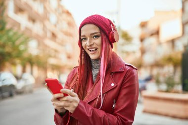 Beyaz kız şehirde akıllı telefon ve kulaklık kullanarak mutlu bir şekilde gülümsüyor..