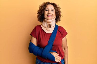Beautiful middle age mature woman wearing cervical collar and arm on sling looking positive and happy standing and smiling with a confident smile showing teeth 