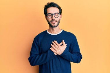 Gündelik kıyafetler ve gözlük takan genç İspanyol adam elleri göğsünde, gözleri kapalı ve yüzünde minnet dolu bir jest ile gülümsüyor. sağlık kavramı. 