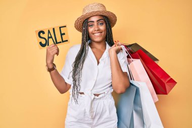 Saçları örgü örgülü, elinde alışveriş poşetleri olan ve yüzünde mutlu ve havalı bir gülümsemeyle gülümseyen genç bir Afro-Amerikan kadını. diş gösterme. 