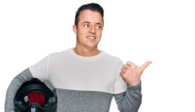 Motosiklet kaskı tutan yakışıklı genç adam başparmağını yana kaldırıyor ve ağzı açık gülümsüyor. 