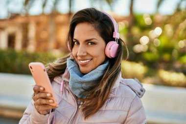 Genç İspanyol kadın şehirde akıllı telefon ve kulaklık kullanarak mutlu bir şekilde gülümsüyor..