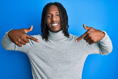 Balıkçı yaka kazak giyen, kendine güvenen, gülümseyen, kendini gururlu ve mutlu parmaklarıyla gösteren Afrikalı Amerikalı bir adam.. 