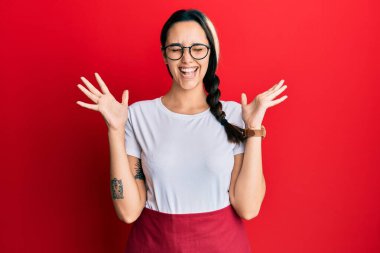 Profesyonel garson önlüğü giyen genç İspanyol kadın başarı için çıldırıyor ve çıldırıyor kollarını kaldırıyor ve gözleri kapalı heyecanla bağırıyor. Kazanan konsept 