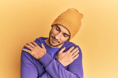 Young handsome man wearing wool winter hat hugging oneself happy and positive, smiling confident. self love and self care 