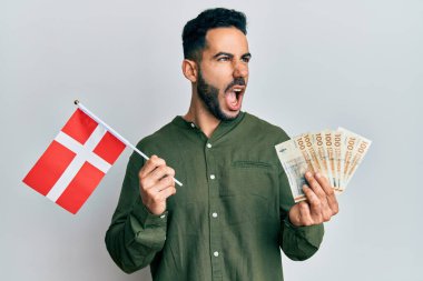 Danimarka bayrağı ve Krone banknotları tutan genç İspanyol adam kızgın ve öfkeli çığlıklar atıyor, öfkeyle bağırıyor. öfke ve agresif konsept. 