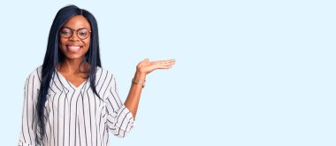 Genç Afrikalı Amerikalı kadın günlük kıyafetler ve gözlük takıyor neşeli bir şekilde gülümsüyor ve avuç içi kameraya bakıyor.. 