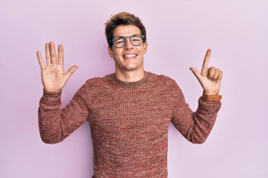 Yakışıklı beyaz bir adam günlük süveter giyiyor ve gözlük takıyor. Yedi numaralı parmaklarını gösterip işaret ediyor. Kendinden emin ve mutlu bir şekilde gülümsüyor.. 