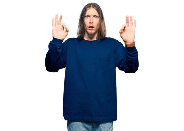 Uzun saçlı, sıradan bir süveter giyen yakışıklı beyaz adam parmaklarıyla onay sembolü yaparken şaşırmış ve şok olmuştu. çılgın ifade 