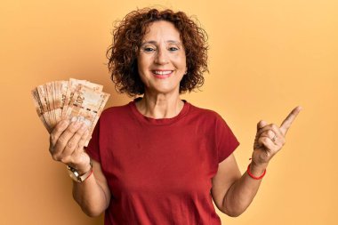 Beautiful middle age mature woman holding south african 20 rand banknotes smiling happy pointing with hand and finger to the side 
