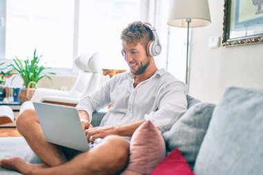 Yakışıklı beyaz adam koltukta oturup bilgisayar kullanarak mutlu mesut gülümsüyor ve kulaklık takarak müzik dinliyor.