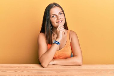 Gündelik giysiler giymiş genç beyaz bir kadın masaya oturmuş kameraya kendinden emin bir şekilde bakıyor kollarını kavuşturmuş ve çenesini kaldırmış gülümsüyor. Olumlu düşünmek. 