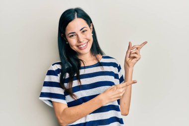 Güzel, genç bir kadın, günlük kıyafetler giyip gülümsüyor ve kameraya bakıyor. İki eliyle ve parmaklarıyla onu işaret ediyor.. 