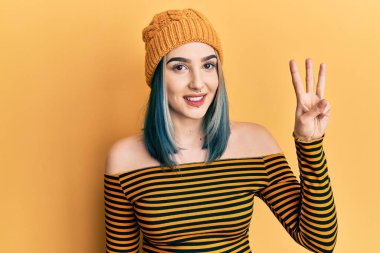 Young modern girl wearing wool hat showing and pointing up with fingers number three while smiling confident and happy. 