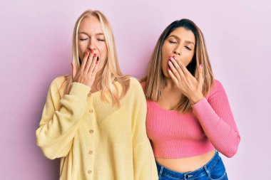 Two friends standing together over pink background bored yawning tired covering mouth with hand. restless and sleepiness. 