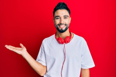 Sakallı genç adam kulaklıkla müzik dinliyor neşeli bir şekilde sunum yapıyor ve avuç içi kameraya bakıyor.. 