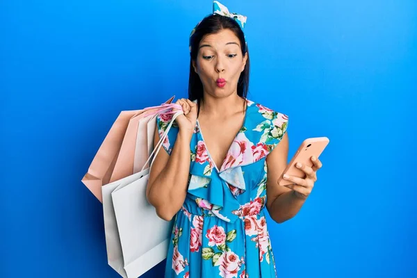 Young latin woman holding shopping bags and smartphone making fish face with mouth and squinting eyes, crazy and comical.